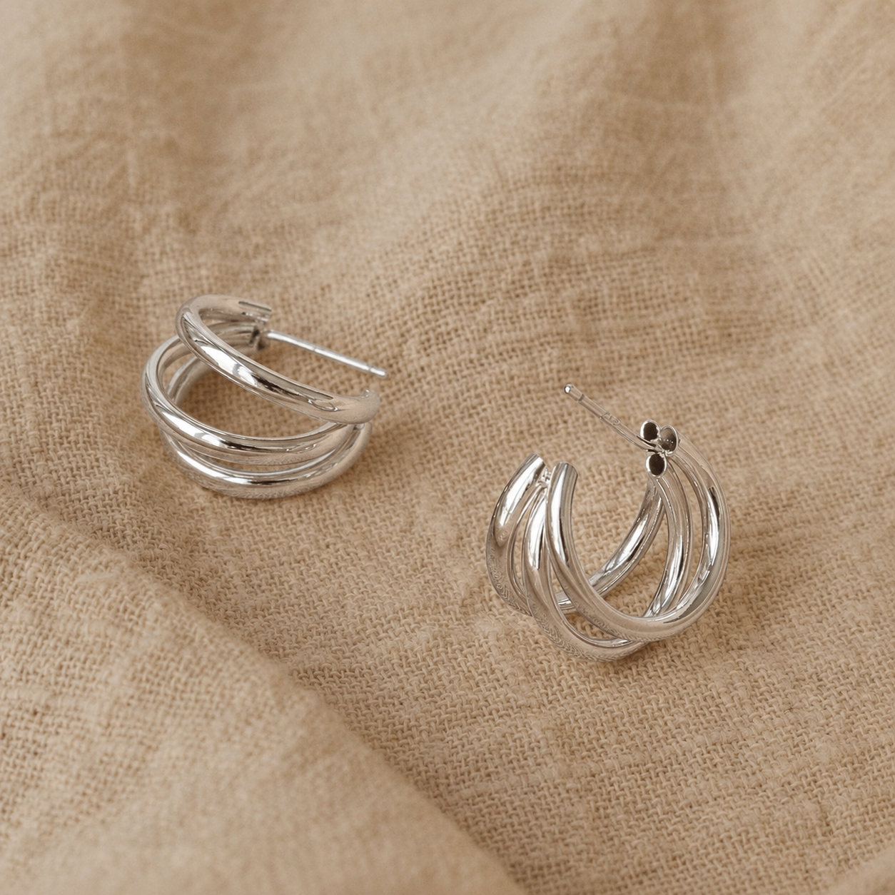 A pair of silver triple-band hoop earrings resting on a textured beige linen fabric background.